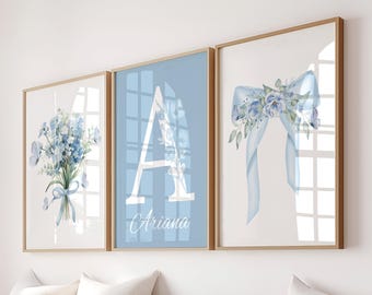 May include: Three framed wall art prints with a floral theme. The prints feature a bouquet of blue flowers, a large letter "A" with the name "Ariana", and a blue bow with floral accents. The frames have a light brown finish.
