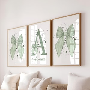 May include: Three framed art prints with a light green bow design. The central print features the letter 'A', a smaller bow, and the name 'Ariana'. Each print has a light background with dark green splatters, all framed in light wood.