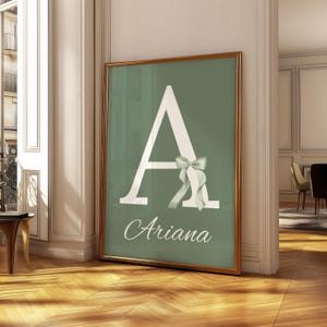Może przedstawiać: Oprawiony wydruk z dużą białą literą "A" na szałwiowym zielonym tle. Do litery przymocowana jest jasnozielona kokarda. Imię "Ariana" jest napisane kursywą poniżej. Rama jest złota.
