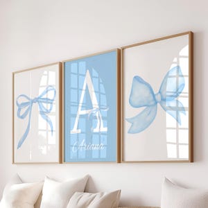 Puede incluir: Tres impresiones artísticas enmarcadas con lazos de acuarela y la letra «A». La impresión central tiene un fondo azul claro con una «A» blanca y el nombre «Ariana». Las otras dos impresiones muestran lazos azul claro sobre un fondo blanco.