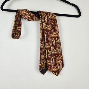 May include: A burgundy tie with a repeating paisley pattern in shades of gold and black. The tie is hanging on a black hanger against a white background. The tie is made of a textured fabric.