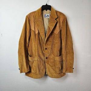 Puede incluir: Blazer de pana marrón claro con solapas de muesca, dos bolsillos delanteros y cierre de dos botones. La chaqueta tiene un detalle de canesú de estilo occidental y una etiqueta dentro del cuello. El blazer está colgado de una percha negra.