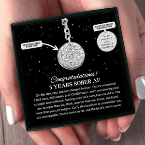 May include: A silver keychain with a circular pendant featuring a star map and a second circular pendant with custom engraving. The black display card reads Congratulations! 5 YEARS SOBER AF and includes text about 1,825 days of sobriety.