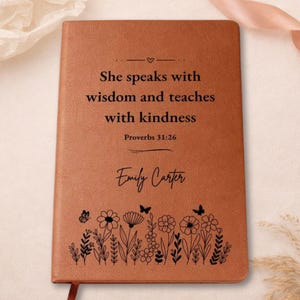 May include: A tan leather-like journal featuring the black printed text "She speaks with wisdom and teaches with kindness Proverbs 31:26 Emily Carter" above a black line art illustration of various wildflowers and small butterflies.