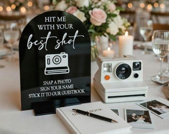 May include: A black sign with white text that reads "HIT ME WITH YOUR best shot" and "SNAP A PHOTO SIGN YOUR NAME STICK IT TO OUR GUESTBOOK". A white Polaroid camera, a guest book, and photos are on a white tablecloth.
