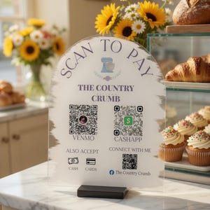 May include: A clear acrylic sign with the text "SCAN TO PAY" and "THE COUNTRY CRUMB". The sign displays QR codes for Venmo and CashApp, along with icons for cash and cards. A black base supports the sign.