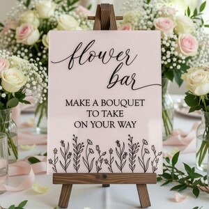 May include: A rectangular light pink acrylic sign displayed on a dark brown wooden easel. The sign features the black text "flower bar" in a cursive script, followed by "MAKE A BOUQUET TO TAKE ON YOUR WAY" and a black line drawing of wildflowers.