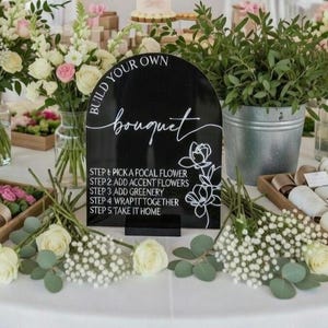 May include: A black arched acrylic sign with white text that reads BUILD YOUR OWN bouquet and lists five steps for flower arrangement, accompanied by a white line drawing of flowers, standing on a small black base on a table with flowers.