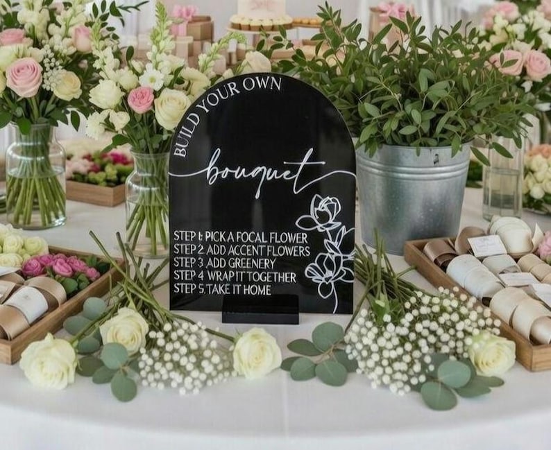 May include: A black arched acrylic sign with white text that reads BUILD YOUR OWN bouquet and lists five steps for flower arrangement, accompanied by a white line drawing of flowers, standing on a small black base on a table with flowers.