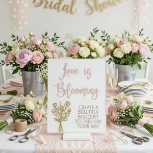 May include: A white rectangular sign with a stand featuring the pink text Love is Blooming and the black text CREATE A BEAUTIFUL BOUQUET TO TAKE ON YOUR WAY, accompanied by a gold line-art illustration of a flower bouquet on the left side.