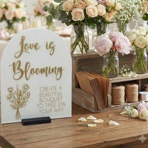 May include: An arched white acrylic sign with gold lettering that reads Love is Blooming and Create a Beautiful Bouquet to Take on Your Way, featuring a gold floral graphic and a black base, displayed on a wooden table with flower arrangements.