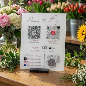 May include: A white rectangular payment sign with the text Scan to Pay, featuring QR codes for Venmo and Cash App, icons for Cash and Apple Pay, and social media handles for bloomsbymichelle, supported by a black base stand on a wooden surface.