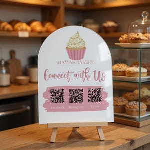 May include: An arched white sign on a wooden easel featuring a cupcake illustration, the text Mama's Bakery, Connect with Us, and three QR codes labeled Facebook, Instagram, and Tiktok over a pink brushstroke graphic on a wooden counter.