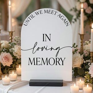 May include: An arched white sign with black text that reads "UNTIL WE MEET AGAIN" in an arc at the top, followed by "IN" in block letters, "loving" in a cursive script, and "MEMORY" in block letters, supported by a black rectangular base.
