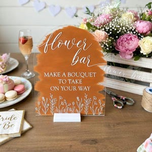 May include: An arched acrylic sign with a burnt orange painted background and white text that reads flower bar MAKE A BOUQUET TO TAKE ON YOUR WAY, featuring white line art of wildflowers at the bottom and a white rectangular base stand.