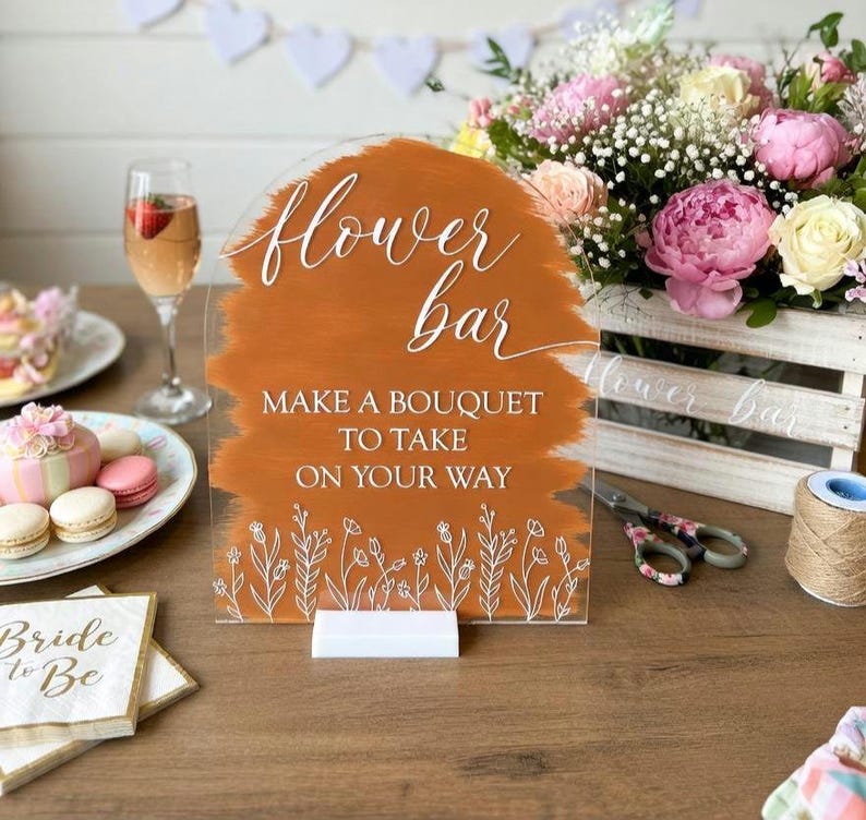 May include: An arched acrylic sign with a burnt orange painted background and white text that reads flower bar MAKE A BOUQUET TO TAKE ON YOUR WAY, featuring white line art of wildflowers at the bottom and a white rectangular base stand.