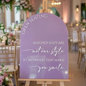 May include: An arched lavender acrylic sign on a wooden easel with white text that reads: OPEN SEATING, ASSIGNED SEATS ARE not our style, SIT ANYWHERE THAT MAKES you smile. The sign features a smooth, solid purple surface with raised lettering.