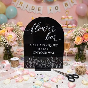 May include: A black arched acrylic sign with white text that reads flower bar, MAKE A BOUQUET TO TAKE ON YOUR WAY, and a line drawing of wildflowers at the bottom. The sign is mounted on a black rectangular base and stands upright.