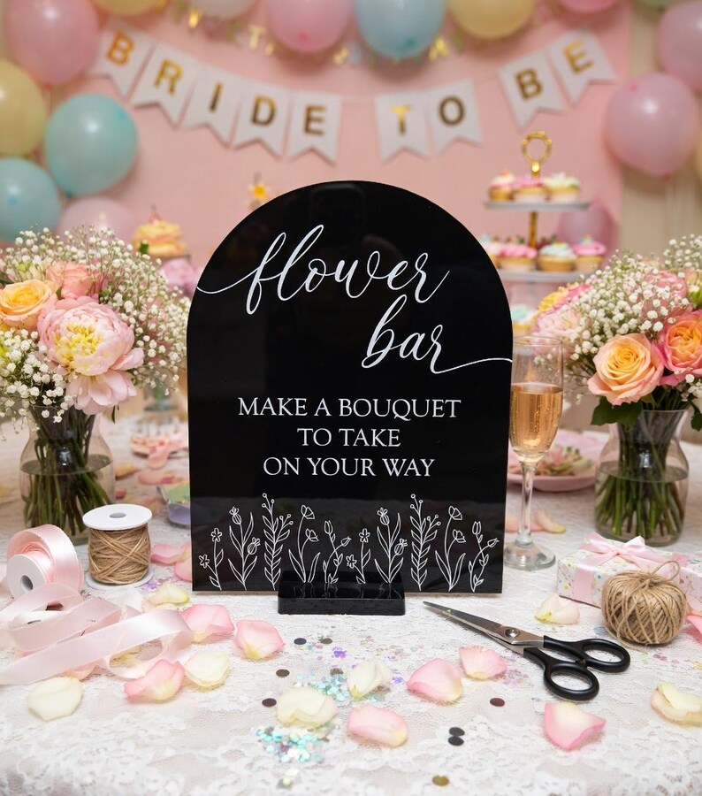 May include: A black arched acrylic sign with white text that reads flower bar, MAKE A BOUQUET TO TAKE ON YOUR WAY, and a line drawing of wildflowers at the bottom. The sign is mounted on a black rectangular base and stands upright.