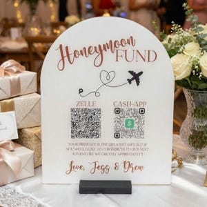 May include: An arched white sign with rose gold text reading Honeymoon Fund, featuring a black airplane icon, Zelle and Cash-App QR codes, and the text Love, Jess & Drew. The sign sits on a black rectangular base on a table with gifts.
