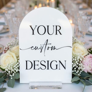 May include: A white arched sign with the words "YOUR custom DESIGN" in black lettering. The sign is displayed on a clear acrylic base, surrounded by white and pink flowers, and greenery. The background features a table setting with glassware and candles.