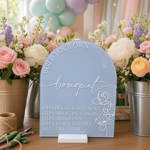 May include: A light blue arched acrylic sign with white text reading BUILD YOUR OWN bouquet and five steps for flower arranging, featuring a white floral line art graphic. The sign is mounted on a white rectangular base on a wooden surface.