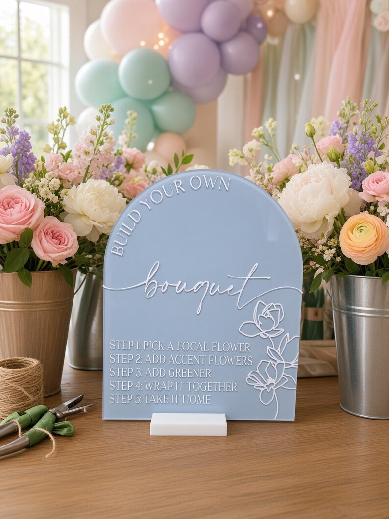 May include: A light blue arched acrylic sign with white text reading BUILD YOUR OWN bouquet and five steps for flower arranging, featuring a white floral line art graphic. The sign is mounted on a white rectangular base on a wooden surface.