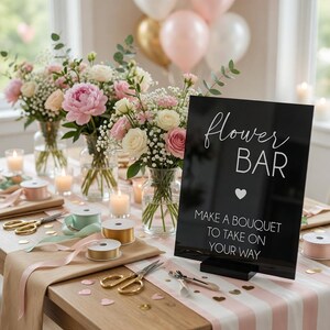 May include: A black rectangular sign with white text reading flower BAR, MAKE A BOUQUET TO TAKE ON YOUR WAY, and a heart icon. The sign sits on a table with glass vases of pink and white flowers, gold scissors, ribbon spools, and candles.