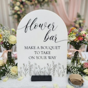 May include: An arched white sign with black text reading flower bar, MAKE A BOUQUET TO TAKE ON YOUR WAY, and a black line drawing of wildflowers at the bottom. The sign is supported by a rectangular black base and features elegant script lettering.