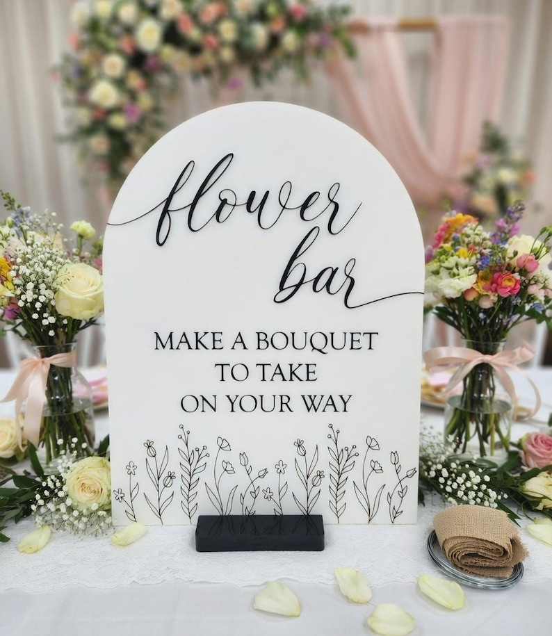 May include: An arched white sign with black text reading flower bar, MAKE A BOUQUET TO TAKE ON YOUR WAY, and a black line drawing of wildflowers at the bottom. The sign is supported by a rectangular black base and features elegant script lettering.