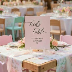 May include: A rectangular light pink sign with black cursive text that reads kids Table and printed text that reads PLEASE HELP YOURSELF with a small heart symbol. The sign is displayed on a table covered with a floral patterned tablecloth.