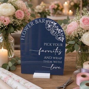 May include: A dark blue arched acrylic sign with white text that reads FLOWER BAR, PICK YOUR favorites, AND WRAP THEM WITH love, featuring a white floral line art design in the top right corner, supported by a rectangular white base.