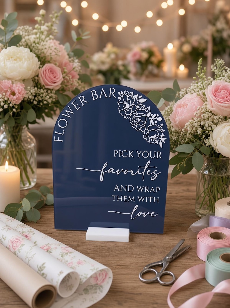 May include: A dark blue arched acrylic sign with white text that reads FLOWER BAR, PICK YOUR favorites, AND WRAP THEM WITH love, featuring a white floral line art design in the top right corner, supported by a rectangular white base.