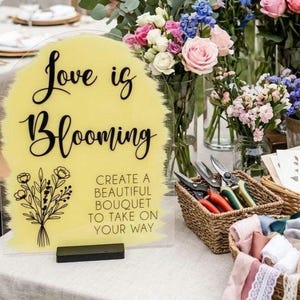 May include: An arched acrylic sign with a yellow painted background featuring the black text "Love is Blooming" and "CREATE A BEAUTIFUL BOUQUET TO TAKE ON YOUR WAY" alongside a black line drawing of a flower bouquet, set on a black base.