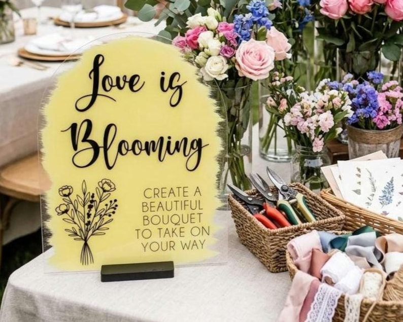 May include: An arched acrylic sign with a yellow painted background featuring the black text "Love is Blooming" and "CREATE A BEAUTIFUL BOUQUET TO TAKE ON YOUR WAY" alongside a black line drawing of a flower bouquet, set on a black base.