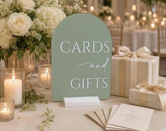 May include: An arched sage green sign with white text that reads CARDS and GIFTS, supported by a rectangular white base. The sign features a clean, modern design with a smooth finish, standing upright as a functional display for event guests.