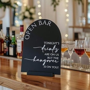 May include: An arched black sign with white text that reads OPEN BAR TONIGHT'S drinks ARE ON US BUT THE hangover IS ON YOU! The sign is mounted on a rectangular black base and features a mix of serif and script fonts for the lettering.