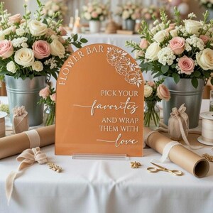May include: An arched terracotta-colored acrylic sign with white text that reads FLOWER BAR PICK YOUR favorites AND WRAP THEM WITH love, featuring a floral line art design in the top right corner, positioned on a white tablecloth with flowers.
