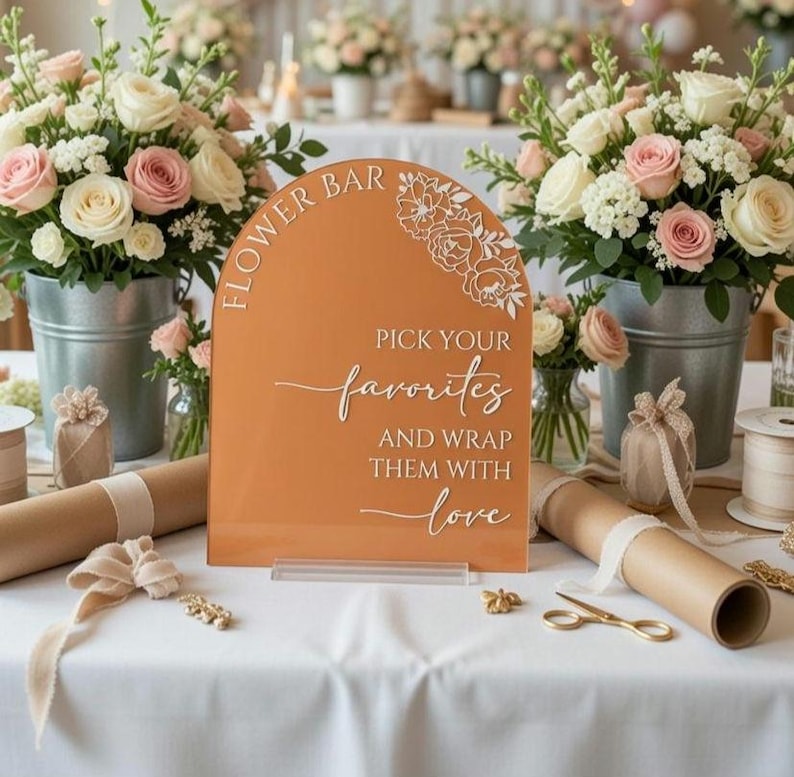 May include: An arched terracotta-colored acrylic sign with white text that reads FLOWER BAR PICK YOUR favorites AND WRAP THEM WITH love, featuring a floral line art design in the top right corner, positioned on a white tablecloth with flowers.