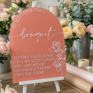 May include: An arched, terracotta-colored acrylic sign with white text reading BUILD YOUR OWN bouquet and five steps for flower arrangement. The sign features a white line-art floral illustration on the right side and rests on a white easel.