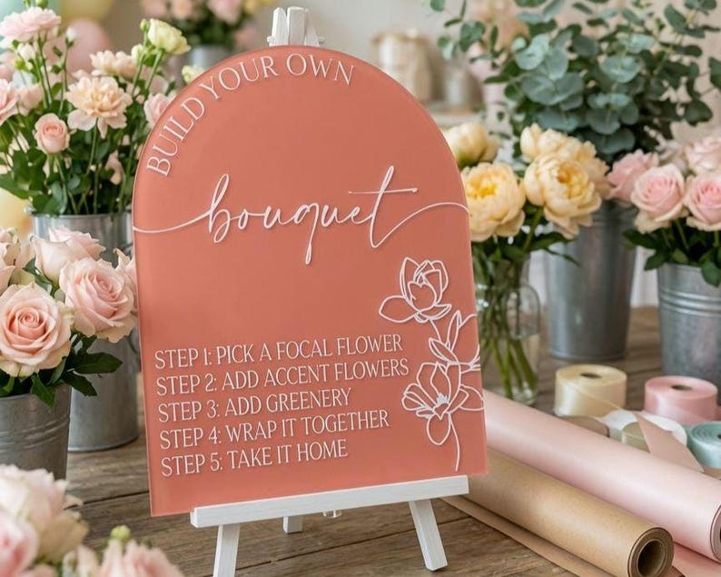 May include: An arched, terracotta-colored acrylic sign with white text reading BUILD YOUR OWN bouquet and five steps for flower arrangement. The sign features a white line-art floral illustration on the right side and rests on a white easel.