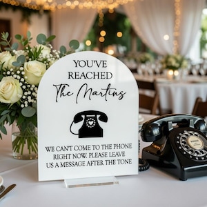 May include: A white acrylic wedding sign with the text "YOU'VE REACHED The Martins" and a phone graphic. A black rotary phone sits next to the sign. The sign reads "WE CAN'T COME TO THE PHONE RIGHT NOW. PLEASE LEAVE US A MESSAGE AFTER THE TONE."