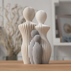 Könnte beinhalten: Eine Familie aus vier Holzskulpturen. Die beiden größeren Figuren sind hellbeige, während die beiden kleineren Figuren grau sind. Jede Figur hat einen abgerundeten Kopf und einen Körper aus vertikalen Holzlatten.