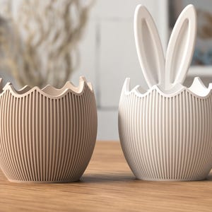 Peut inclure: Deux contenants décoratifs en forme d'œuf avec des rainures verticales. L'un est beige, l'autre est blanc avec des oreilles de lapin. Les contenants ont un bord festonné. Les objets sont sur une surface en bois, suggérant un thème de Pâques.