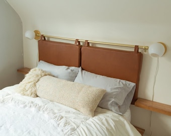 Könnte beinhalten: Ein weißes Bett mit einem braunen Lederkopfteil und einem weißen, flauschigen Kissen. Das Kopfteil ist an einer goldenen Metallstange befestigt. An der Wand befinden sich zwei weiße Nachttischlampen mit goldenen Akzenten.