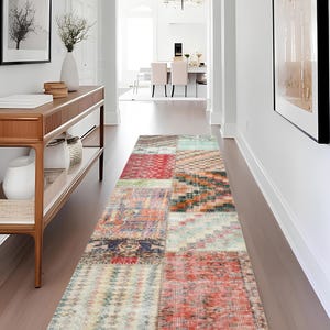 Pode incluir: Um tapete longo e retangular de patchwork com uma variedade de cores e padrões. O tapete apresenta uma mistura de quadrados vermelhos, laranja, azul e bege, criando uma estética vintage ou boêmia. O tapete é colocado em um corredor com paredes claras e piso de madeira.