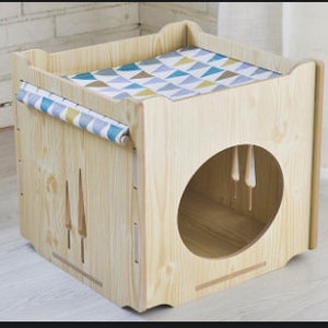 May include: A wooden cat house with a removable cushion on top. The house has a round entrance hole and a small opening on the side. The cushion is covered in a blue and brown geometric pattern.
