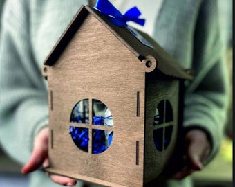 Puede incluir: Una caja de regalo de madera con forma de casa con una cinta azul y una ventana que muestra papel de seda azul en el interior.