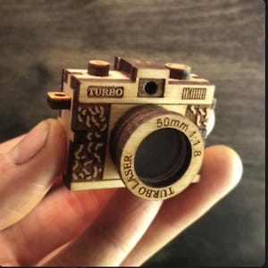 May include: A wooden toy camera with a brown lens and the text "TURBO LASER 50mm 1:1.8" on the lens. The camera is made of wood and has a brown finish.