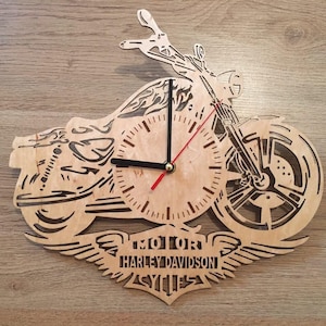 May include: A laser-cut wooden wall clock shaped like a motorcycle, featuring a circular clock face with black hour and minute hands and a red second hand. Below the clock face, a winged emblem displays the text MOTOR HARLEY DAVIDSON CYCLES.